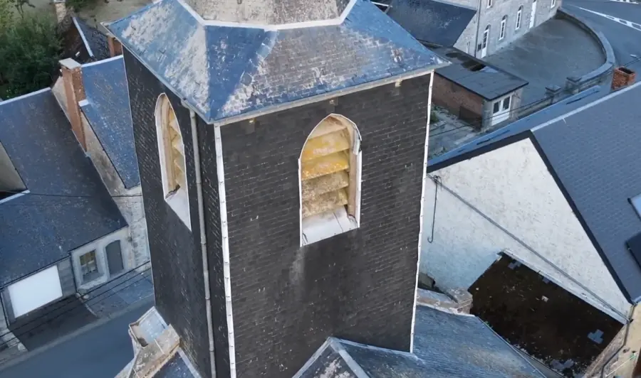 Face d'un beffroi d'église : baie en arc surmontée d'abat-sons en lames de bois, encadrement de pierre, toitures d'ardoises en contrebas.