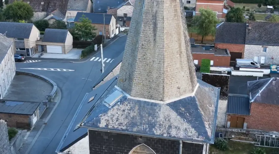 Flèche d'église en ardoises vue depuis le dessus, ses quatre pans convergeant vers la pointe ; rue, maisons et toitures du village tout autour.