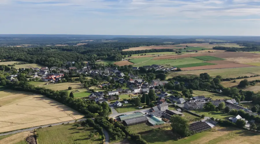 Vue aérienne oblique d'un village entouré de champs et de bosquets, sous un ciel de traîne, l'horizon dégagé au loin.