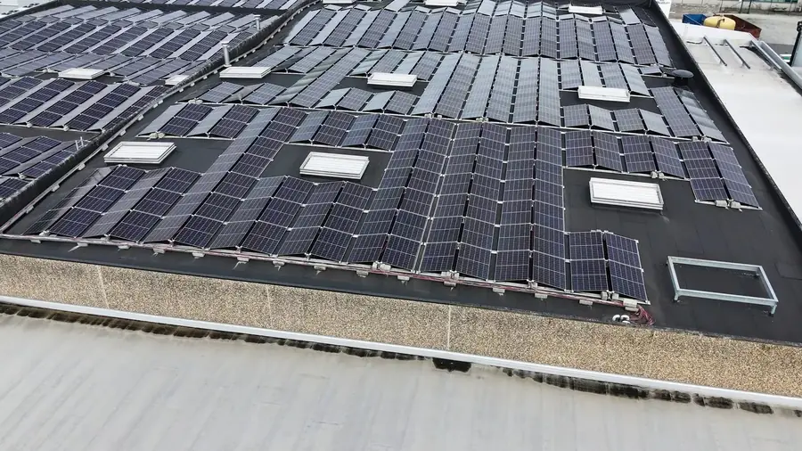 Toiture industrielle plate en membrane bitumineuse entièrement couverte de rangées de modules photovoltaïques inclinés, avec plusieurs lanterneaux blancs intercalés et un acrotère en béton sans garde-corps au premier plan.