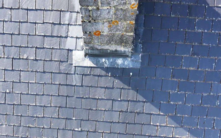 Pied d'une souche de cheminée maçonnée sur un versant d'ardoises neuves ; le solin plomb et l'abergement du raccord sont visibles, la maçonnerie porte des colonies de lichen orange.