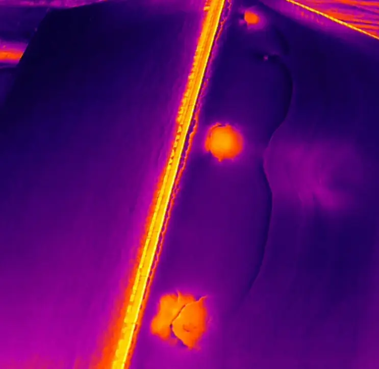 Image infrarouge d'une toiture : fond violet uniforme, arête vive en jaune, deux taches orange nettes et une trace diffuse plus claire.